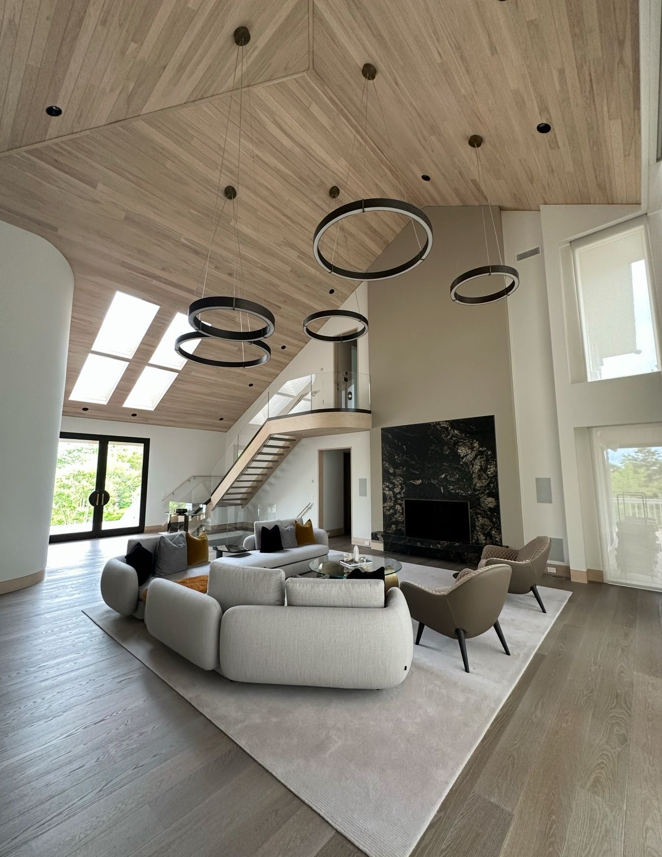 Spacious, modern living room with high ceilings, wood accents, and a large black fireplace. Circular lights hang above a large, light-colored sofa.