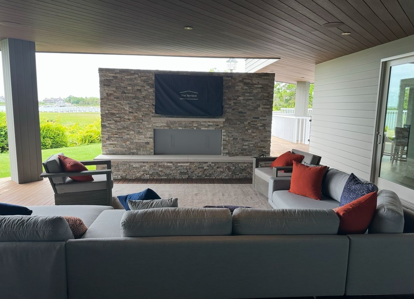 Covered patio with sectional seating and a stone fireplace. Orange and blue pillows accent the gray furniture.