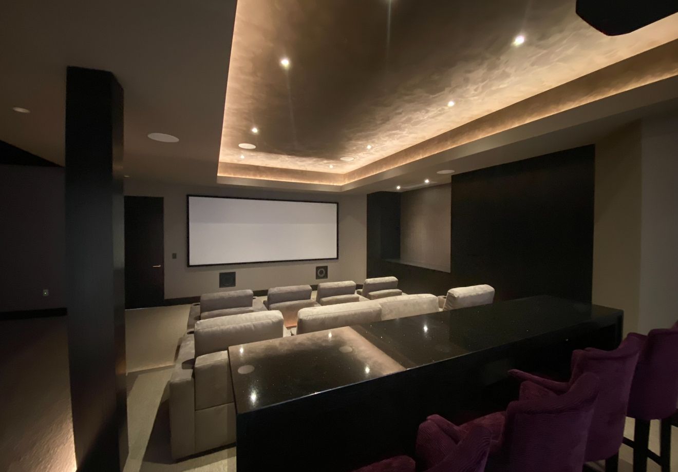 Home theater with a large screen, tiered seating, a black bar, and purple bar stools. The ceiling has recessed lighting.