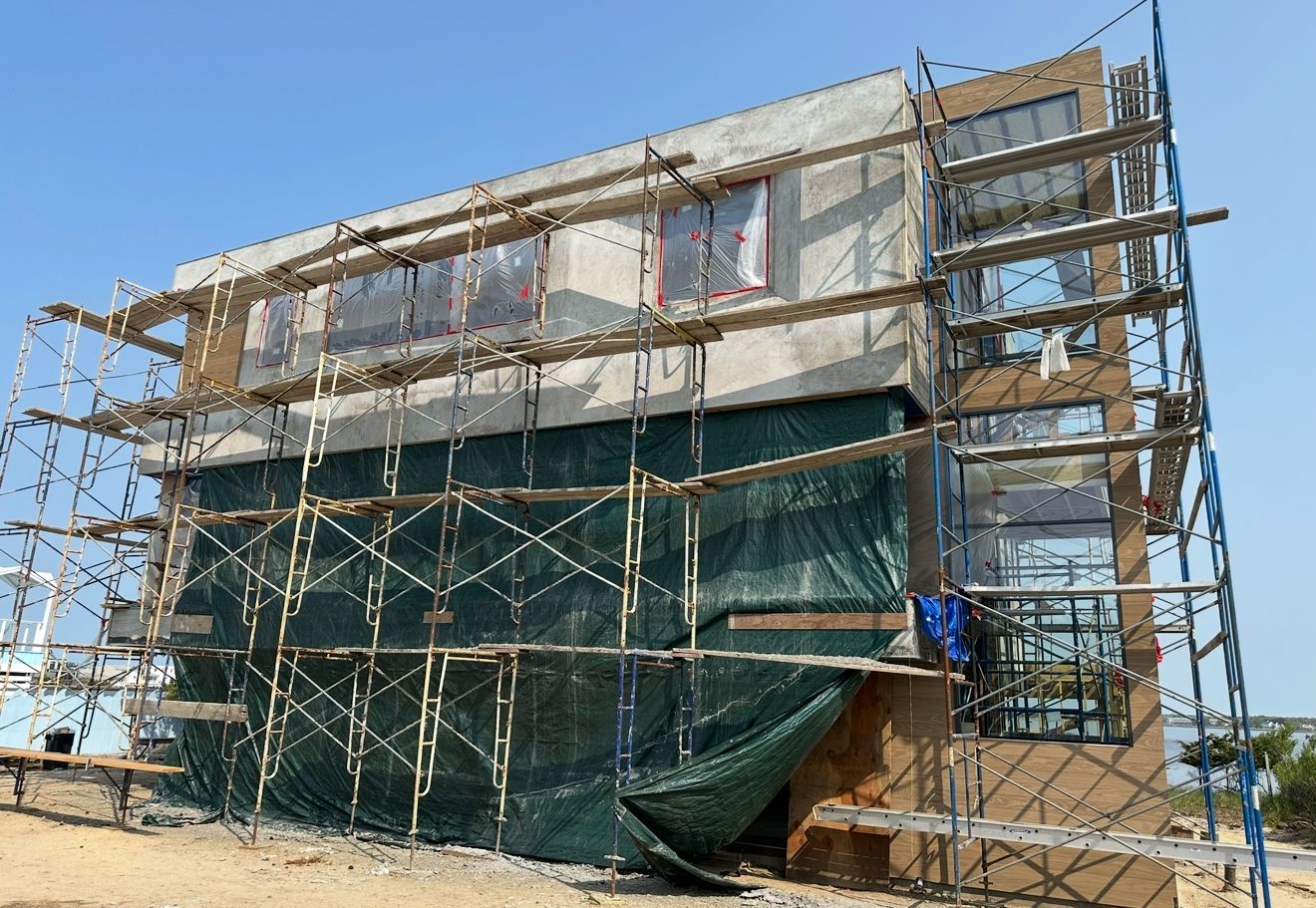 Building under construction with scaffolding, covered in green tarp, against a clear blue sky.