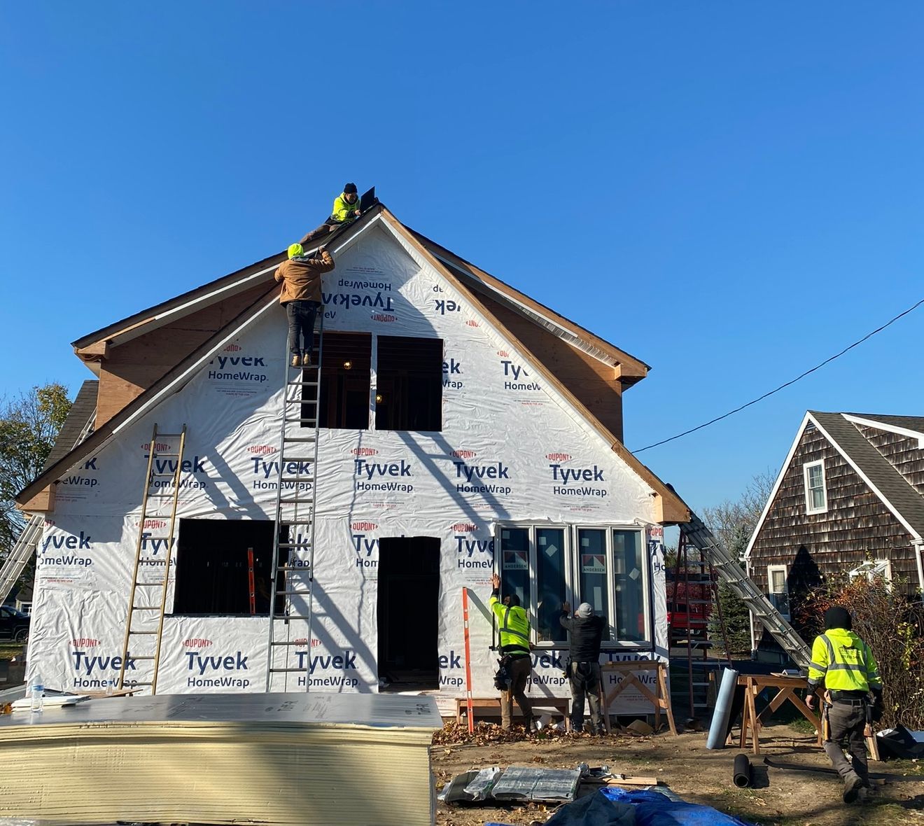 Construction workers installing siding and windows on a two-story house, wrapped in Tyvek. Bright blue sky.