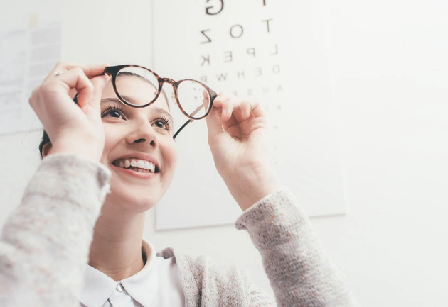Woman holding glasses up in front of an eye chart, smiling in a bright clinic setting