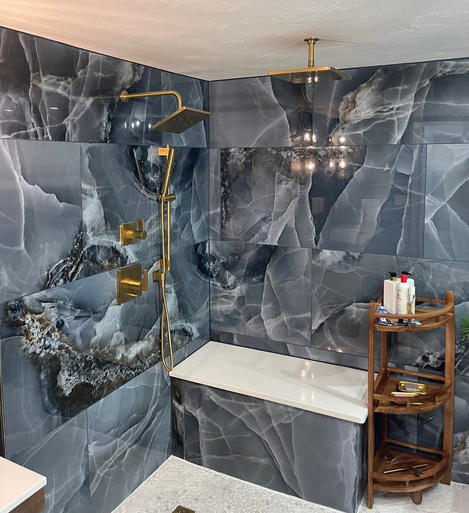 Blue-gray marble bathroom with gold shower fixtures, bench seating, and a small corner shelf.