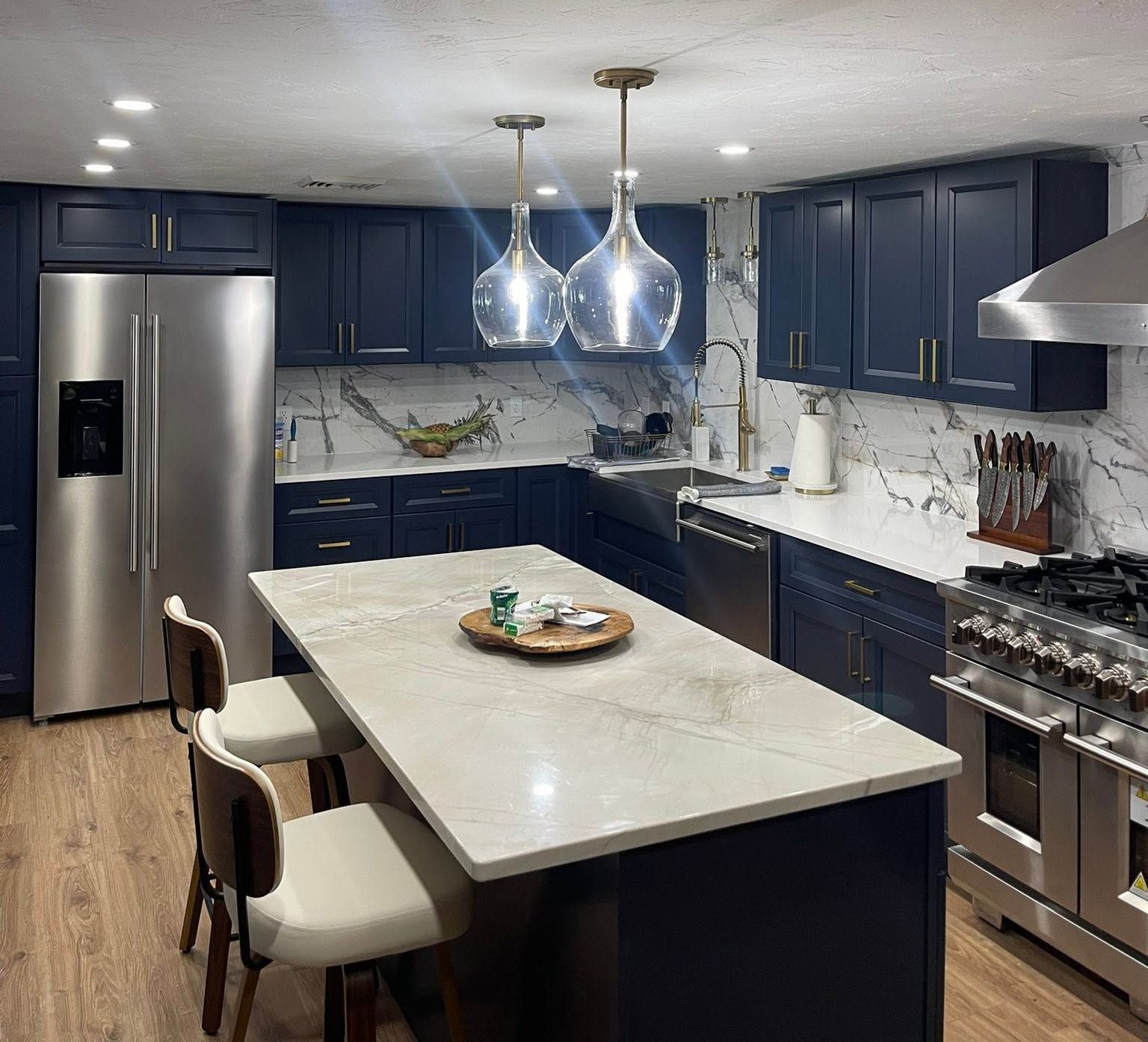 Modern kitchen with navy cabinets, stainless steel appliances, marble island, and pendant lights.