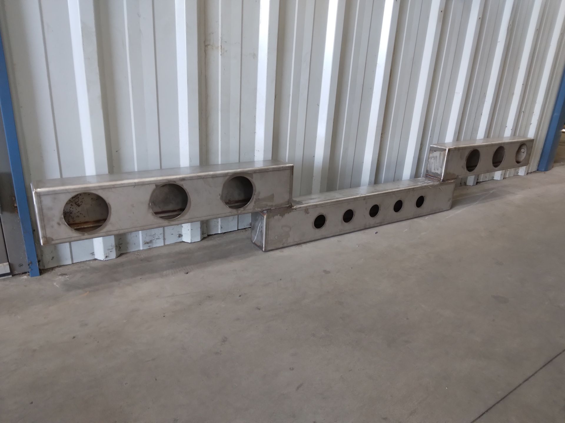 A stainless steel bumper is sitting on the ground in front of a metal wall.