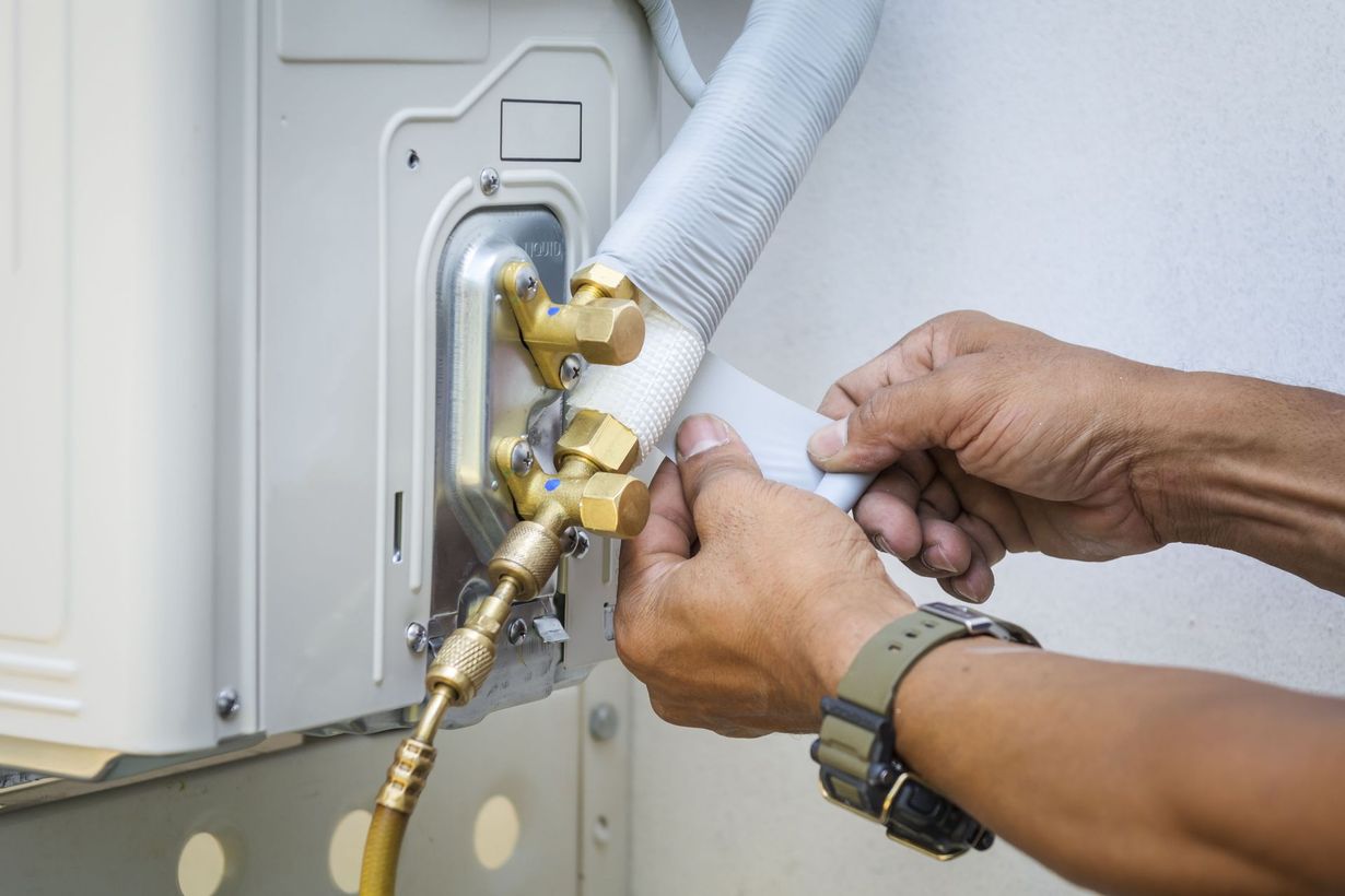 Hands connecting tubing to an outdoor air conditioning unit, showcasing copper fittings and white insulation.