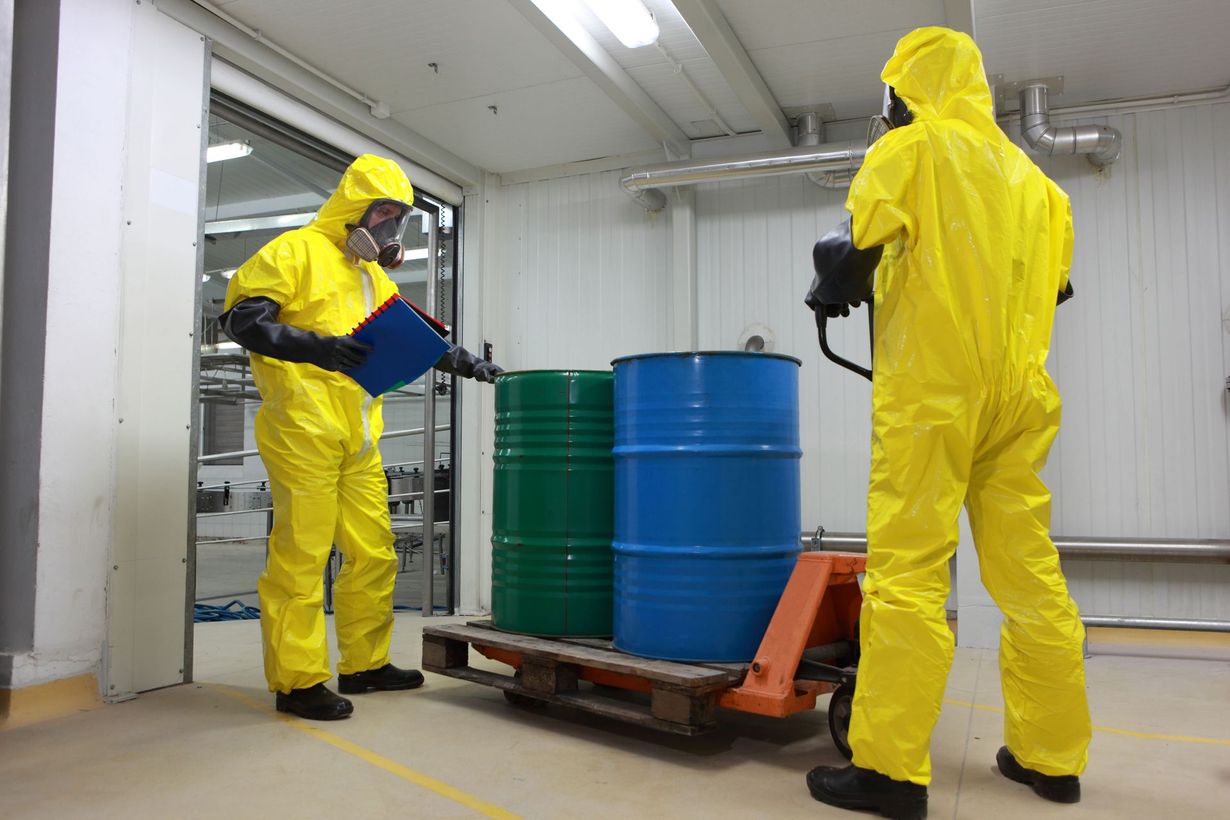 Two people in yellow hazmat suits move barrels on a pallet jack in a lab.