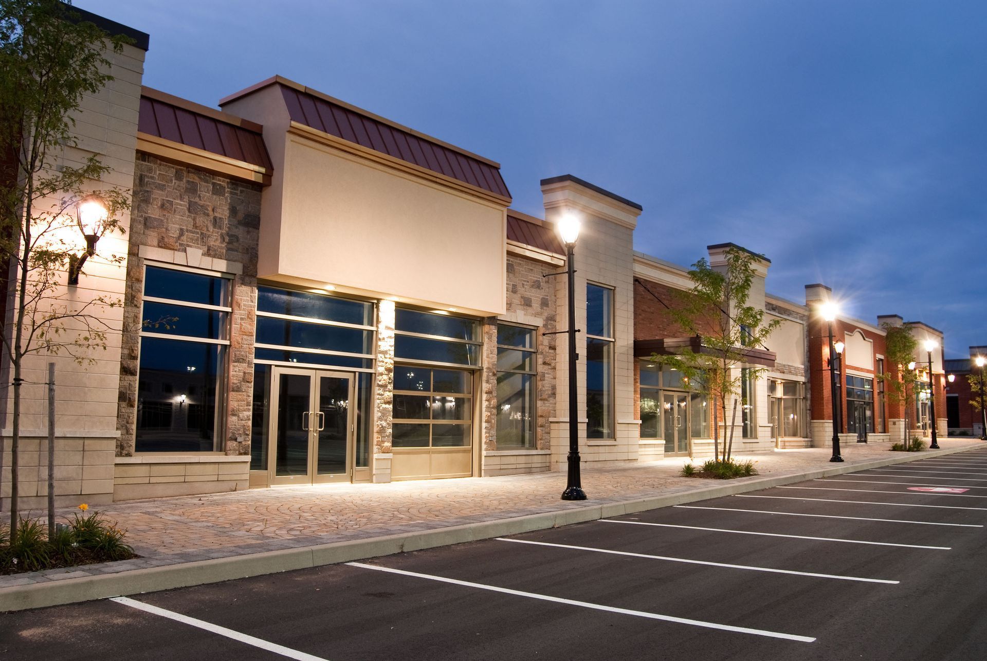 Row of empty commercial storefronts, illuminated at dusk, with brick and tan exteriors and street lights.