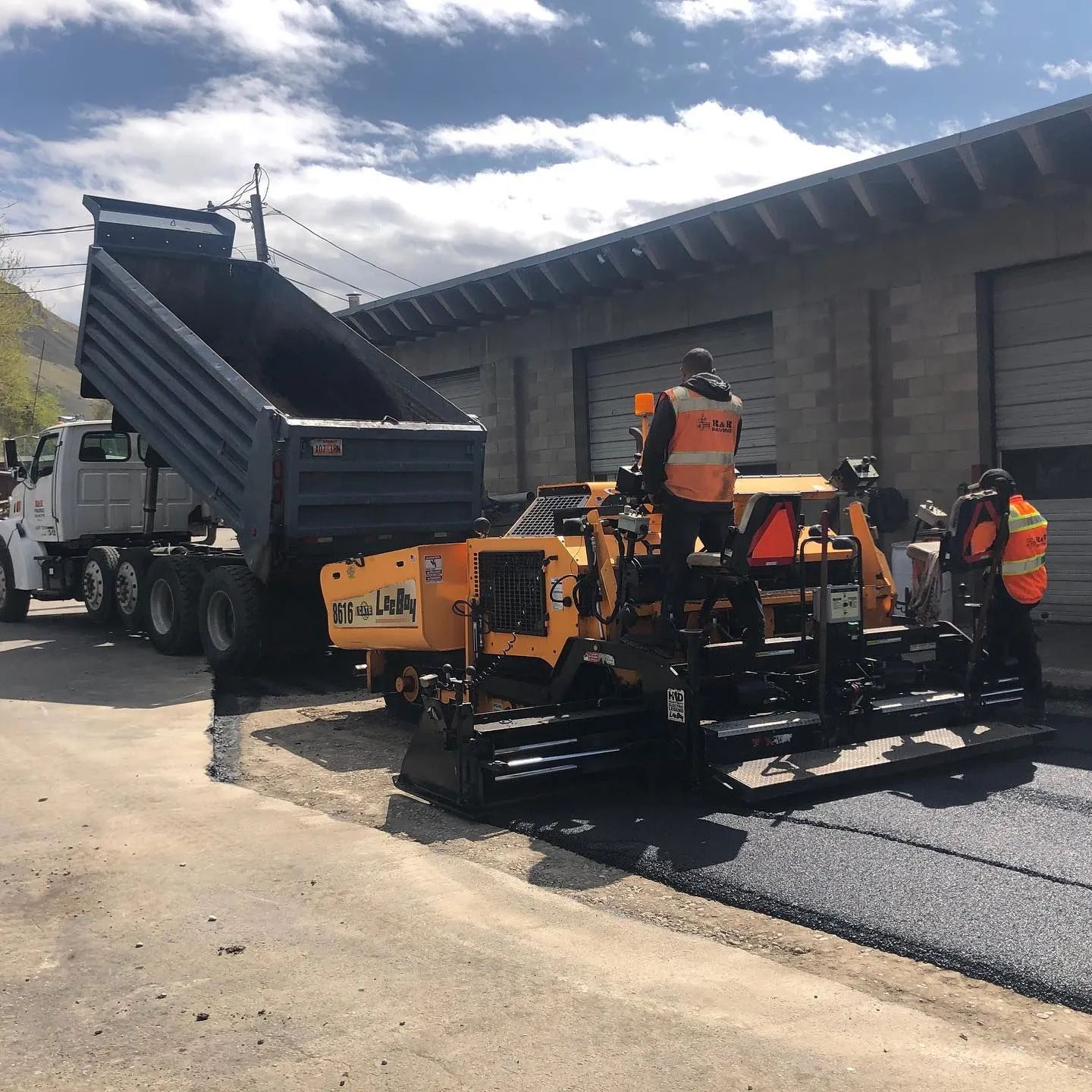 Asphalt paving in progress: Dump truck unloading onto a paver, workers in orange vests.