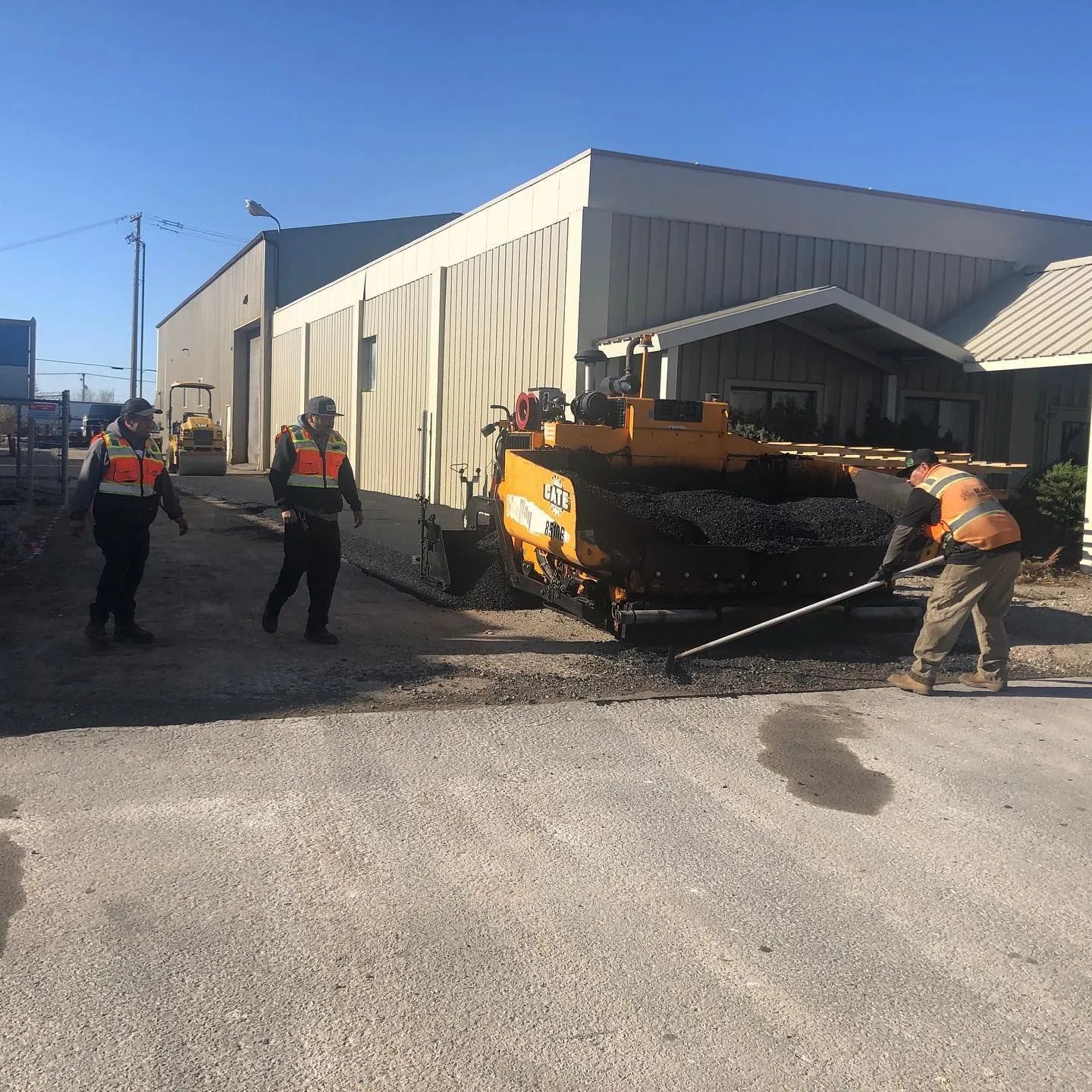 Workers paving asphalt outside a building. One operates machinery, two others assist.