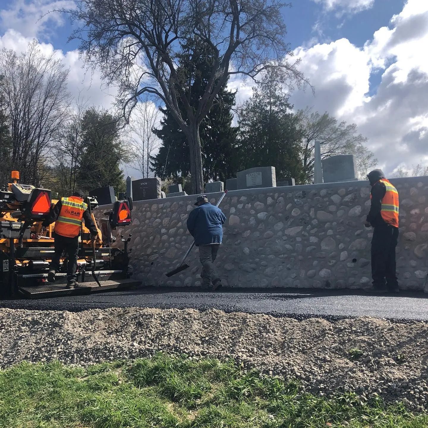 Road crew paving a road next to a stone wall; workers in orange vests.