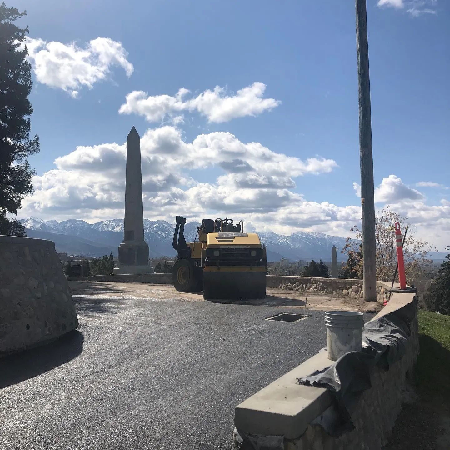 Construction vehicle paving near a tall obelisk with mountains in the background under a blue sky.