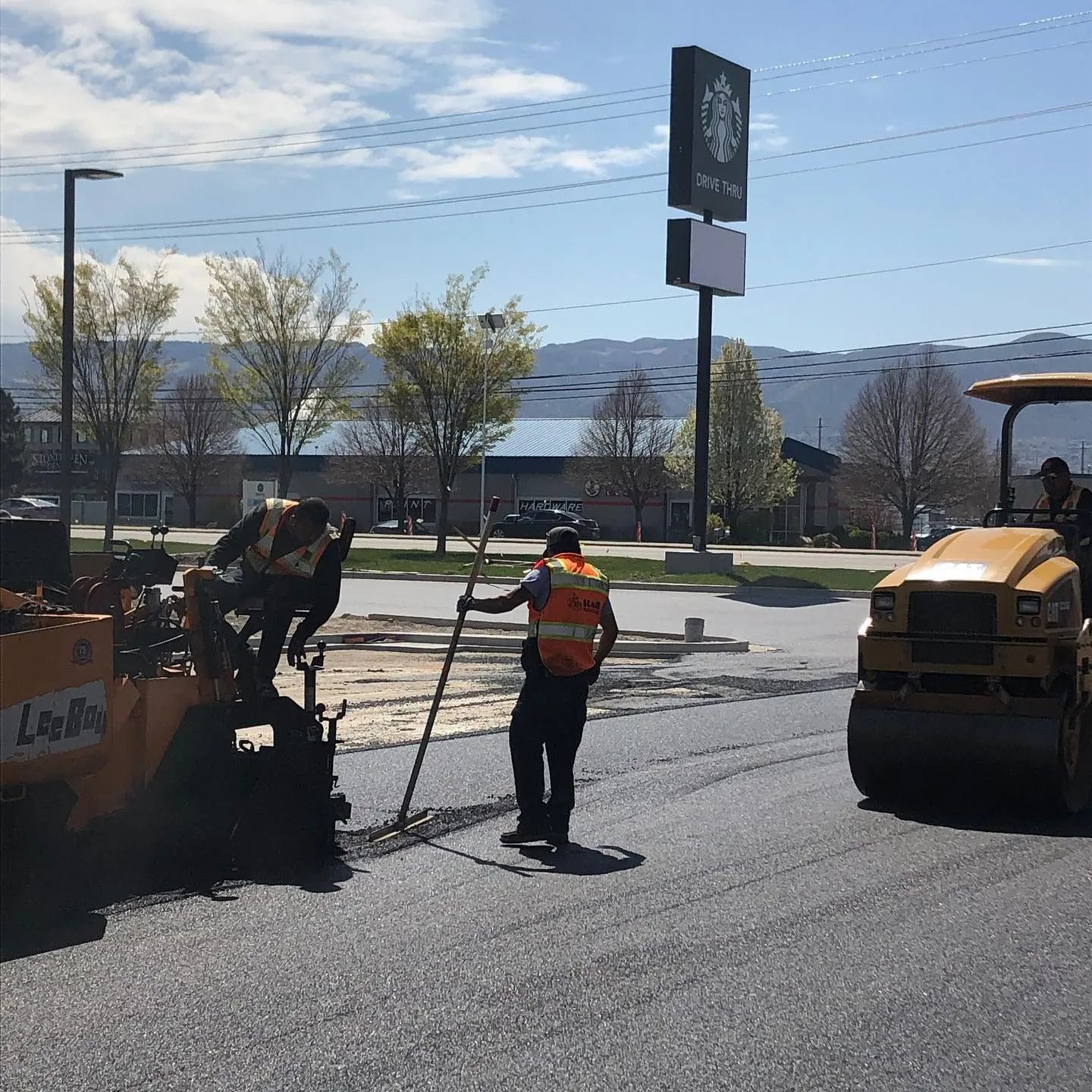 Road crew paving asphalt near a Starbucks, with a paver, roller, and workers.