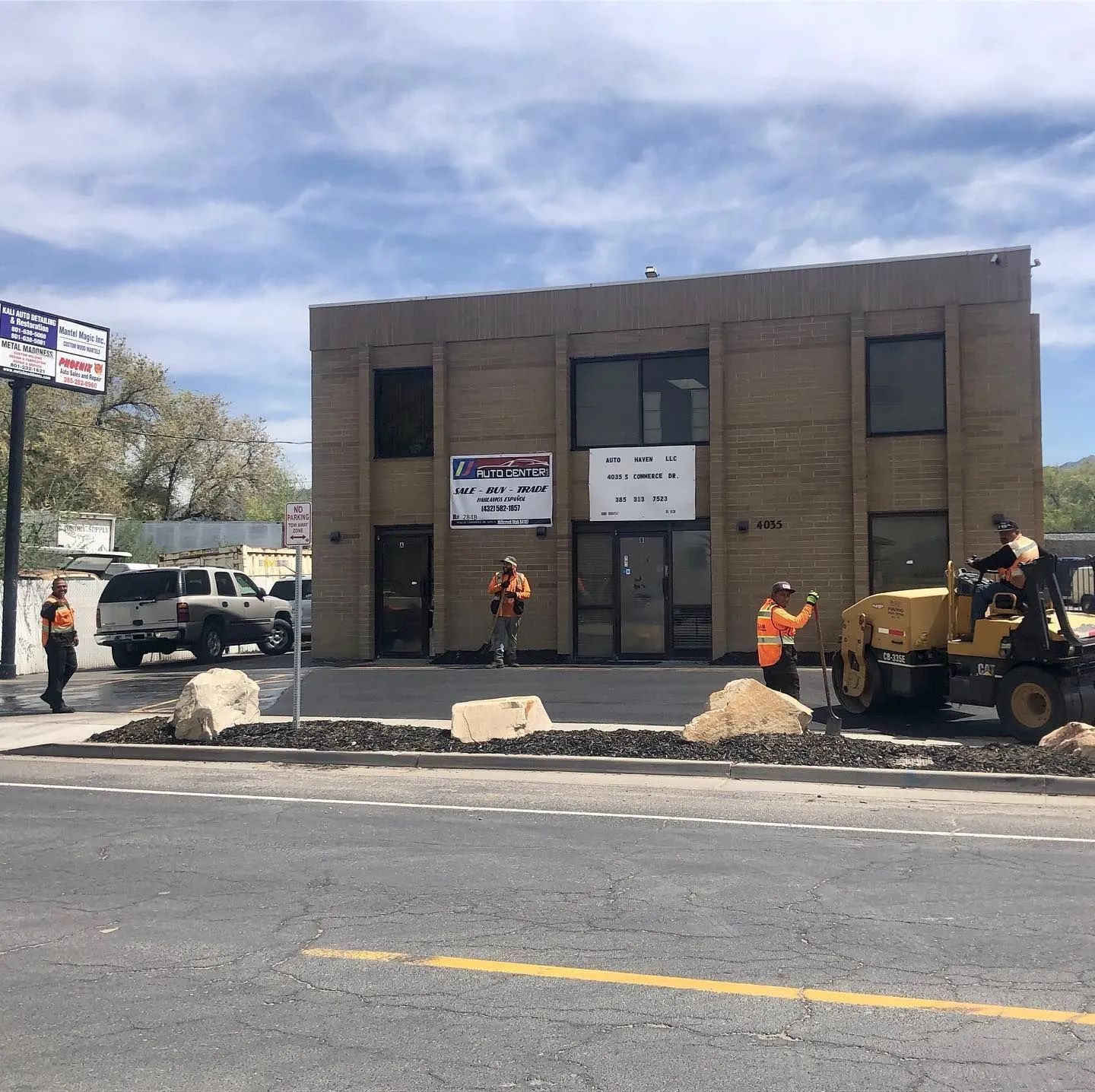 Road construction in progress in front of a tan two-story building; workers in safety vests; heavy machinery.