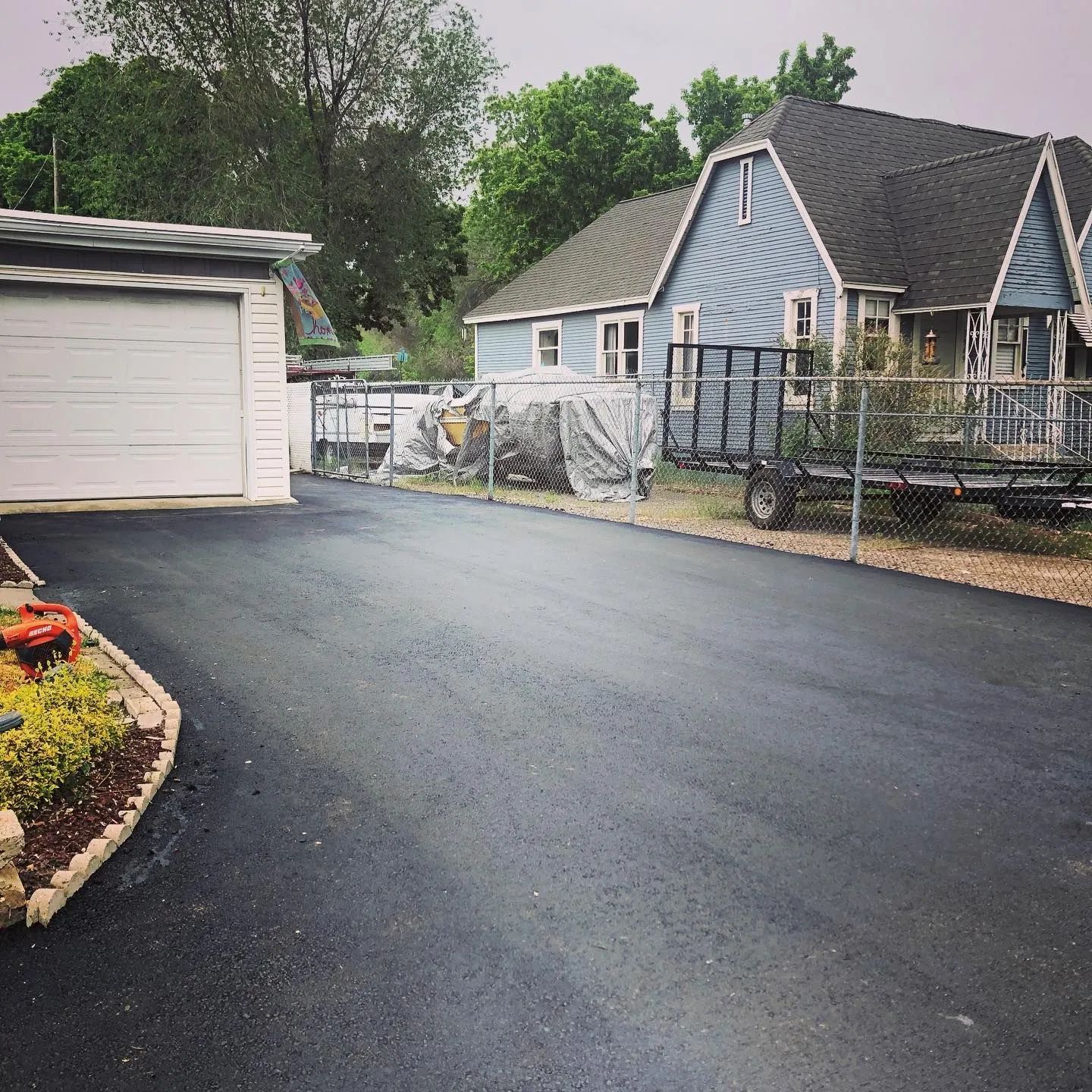 Paved driveway leading to a white garage and blue house with trailer in yard. Green trees in background.