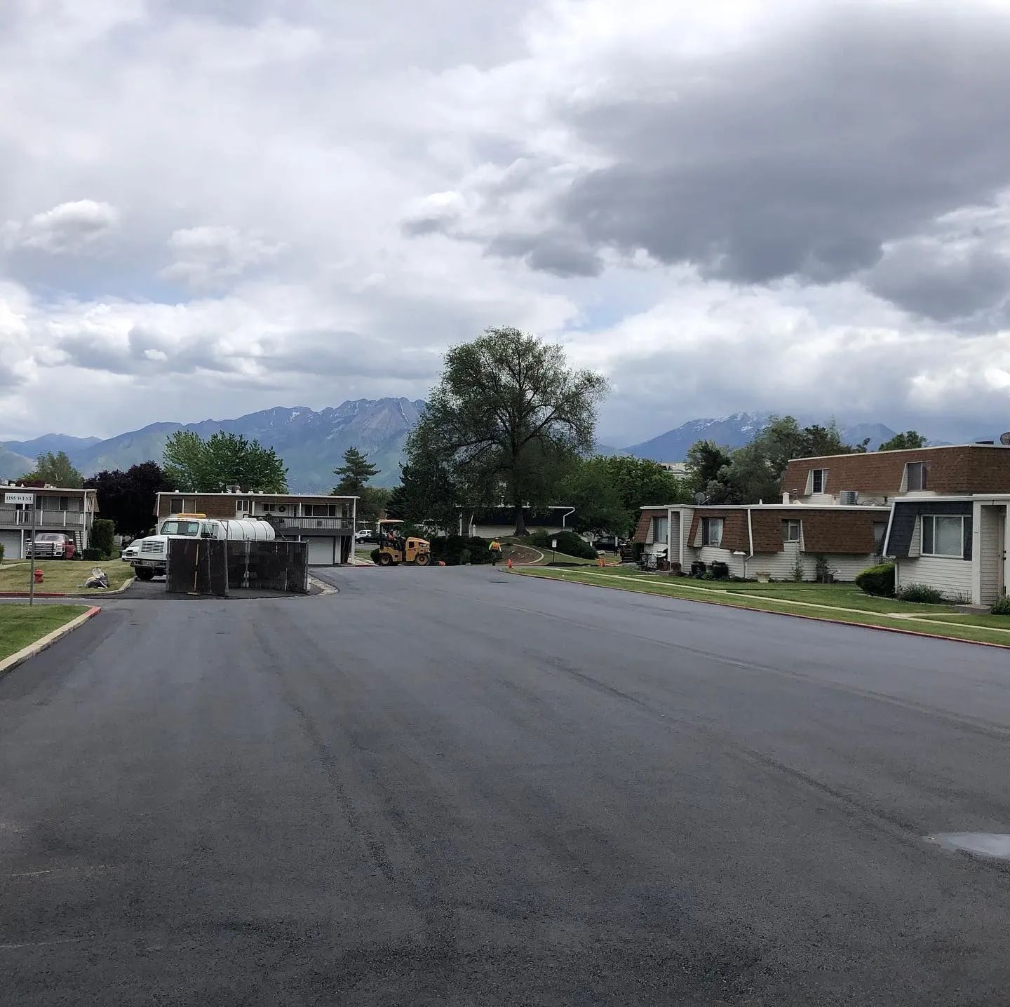 Newly paved black asphalt road in a residential area, with townhouses, trees, and mountains in the distance under a cloudy sky.