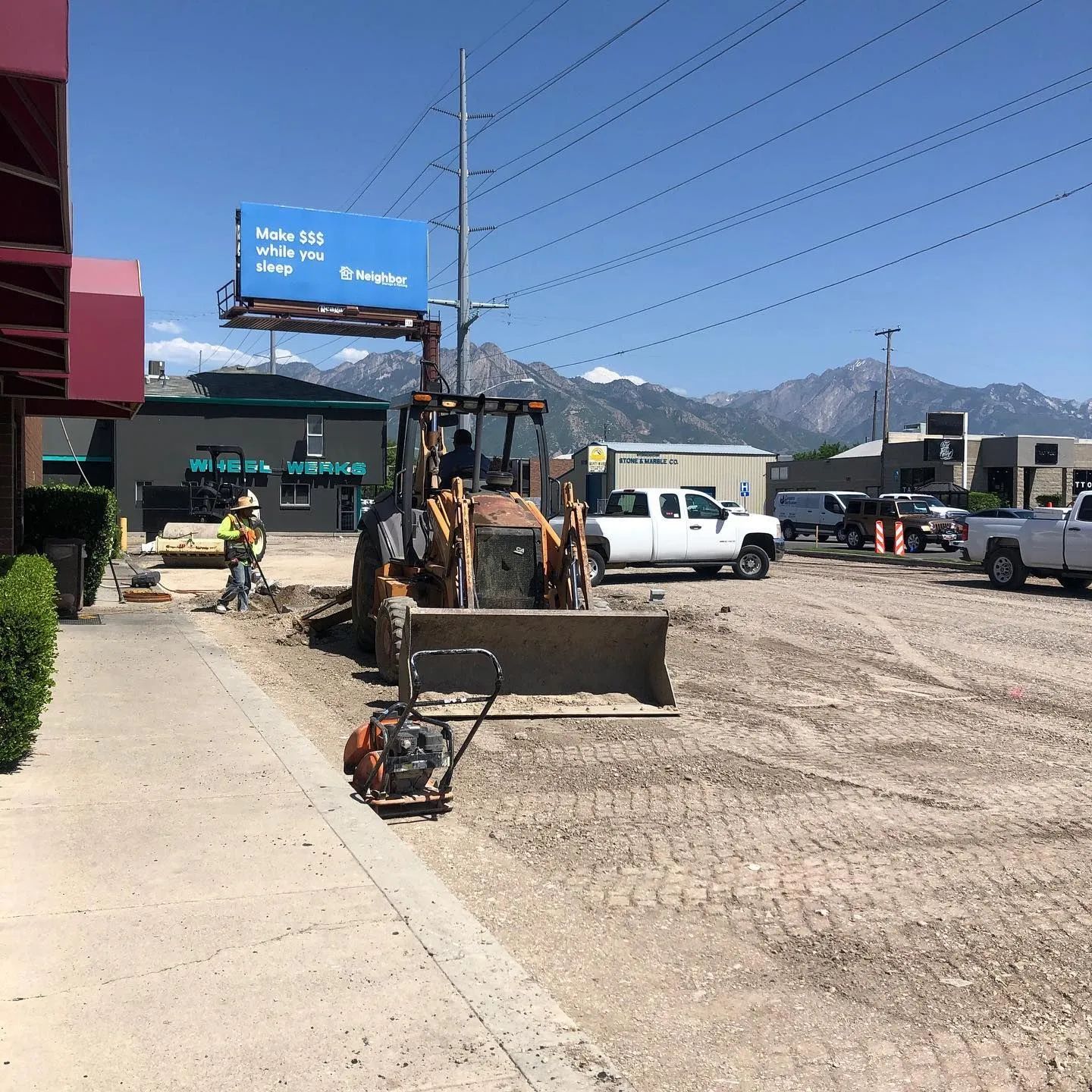 Construction site with backhoe, worker, and parked trucks. Mountains visible in the background. Sunny day.