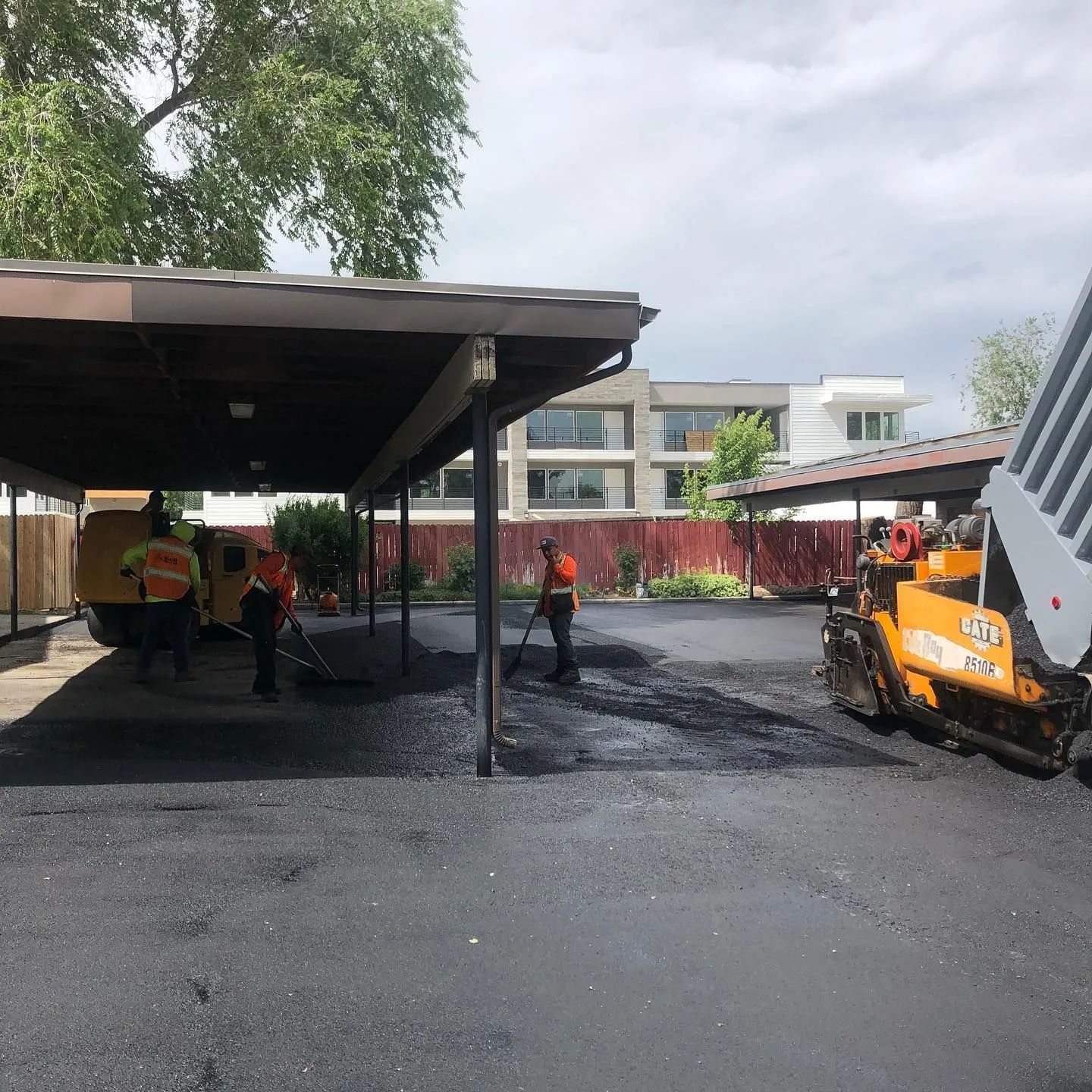 Workers paving a parking lot under a carport. A dump truck pours asphalt, and workers rake it.