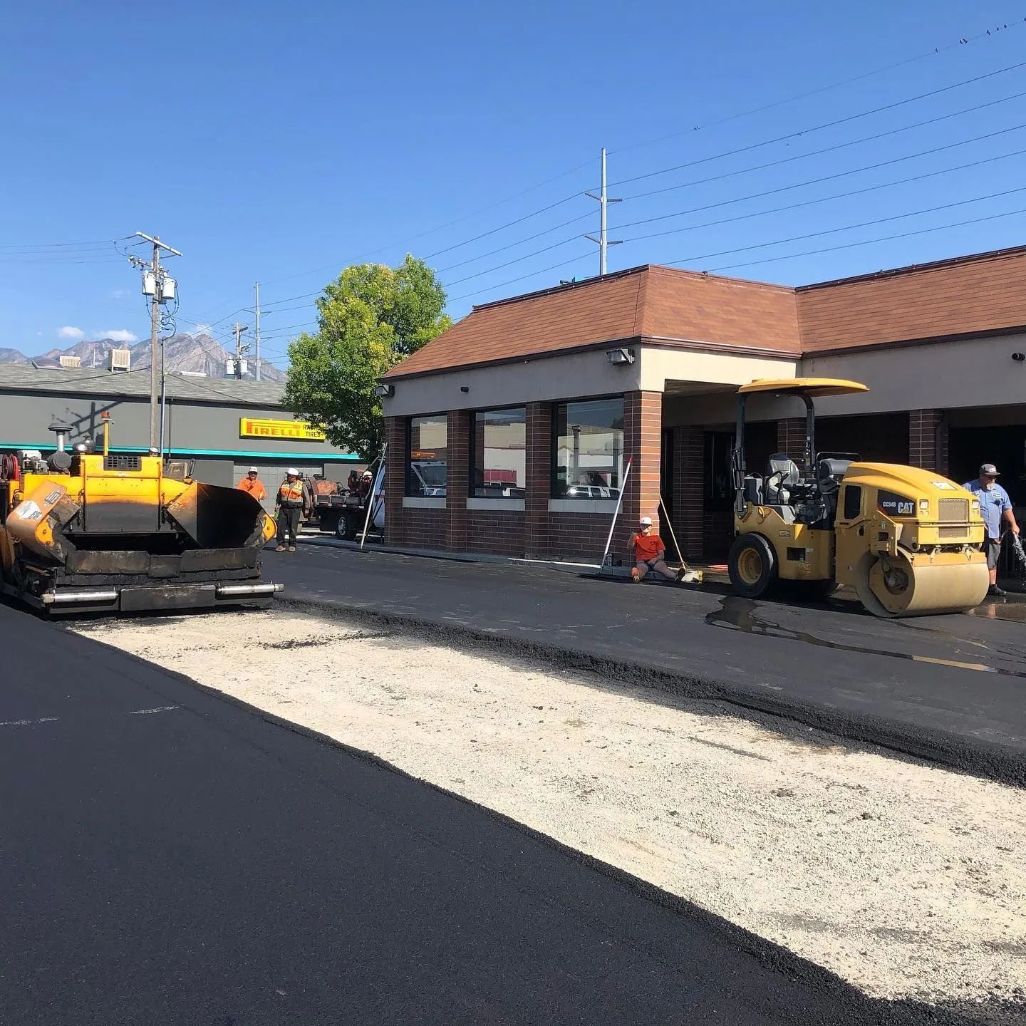 Asphalt paving in progress near a commercial building; yellow machinery, workers, blue sky.