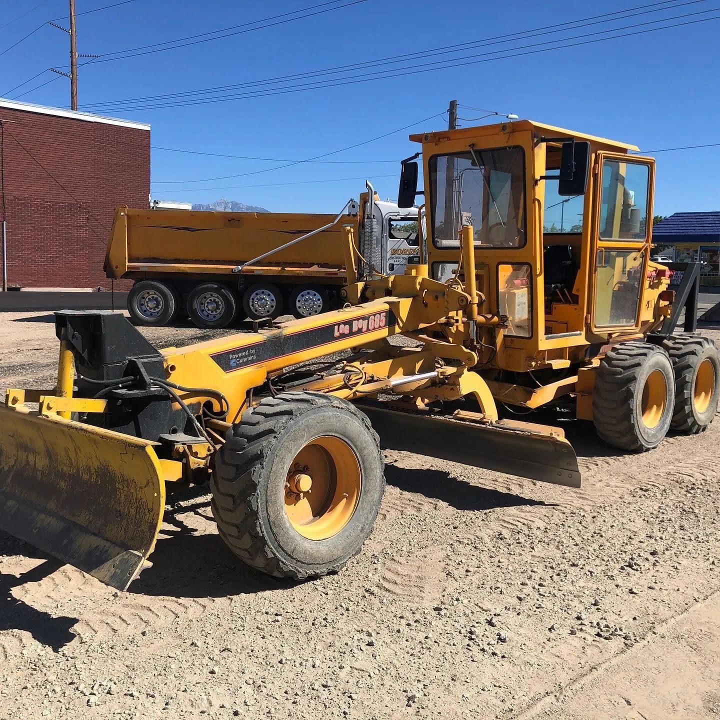 Yellow grader and dump truck on gravel in front of a brick building under a blue sky.