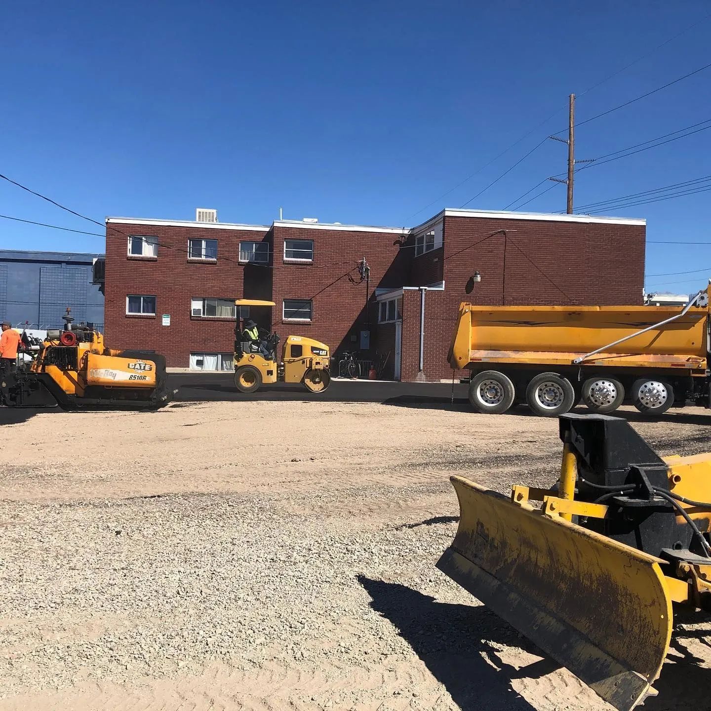 Road construction: Asphalt paving with machinery, including a yellow dump truck and bulldozer, near a brick building.