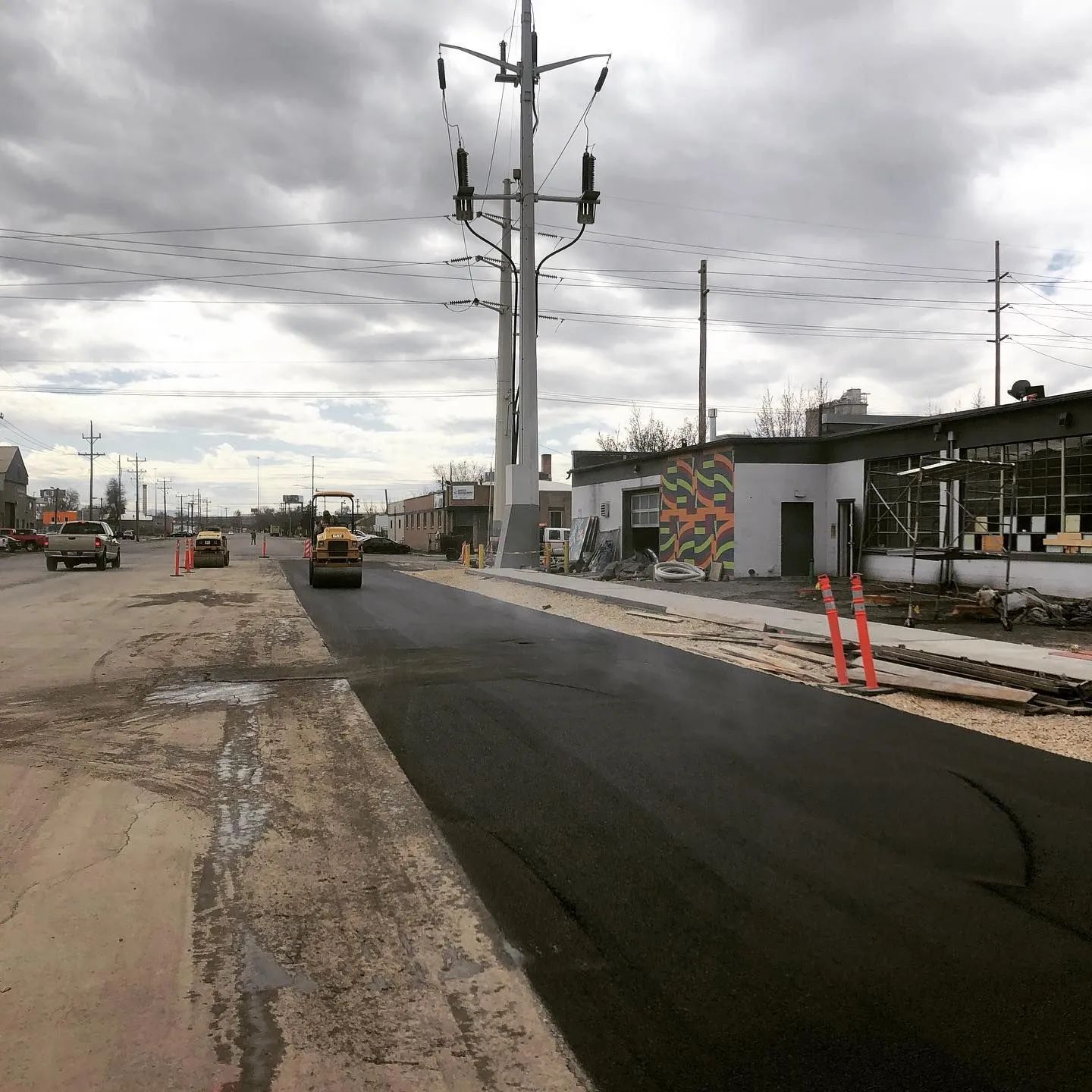 Road paving in progress; asphalt, construction vehicles, power lines, and cloudy sky.