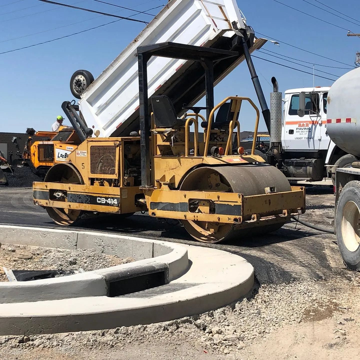 Yellow road roller compacting asphalt near a curb; a dump truck is emptying material nearby.