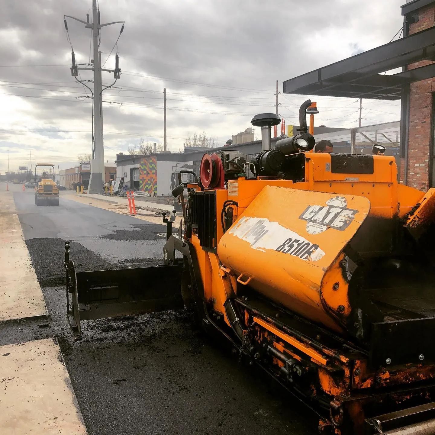 Road paving machine laying asphalt on a street; a roller is in the distance. Cloudy day.