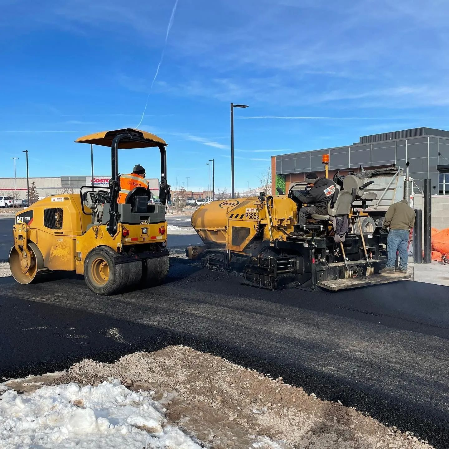 Asphalt paving with a yellow roller and paver machines, workers, and a building on a sunny day.