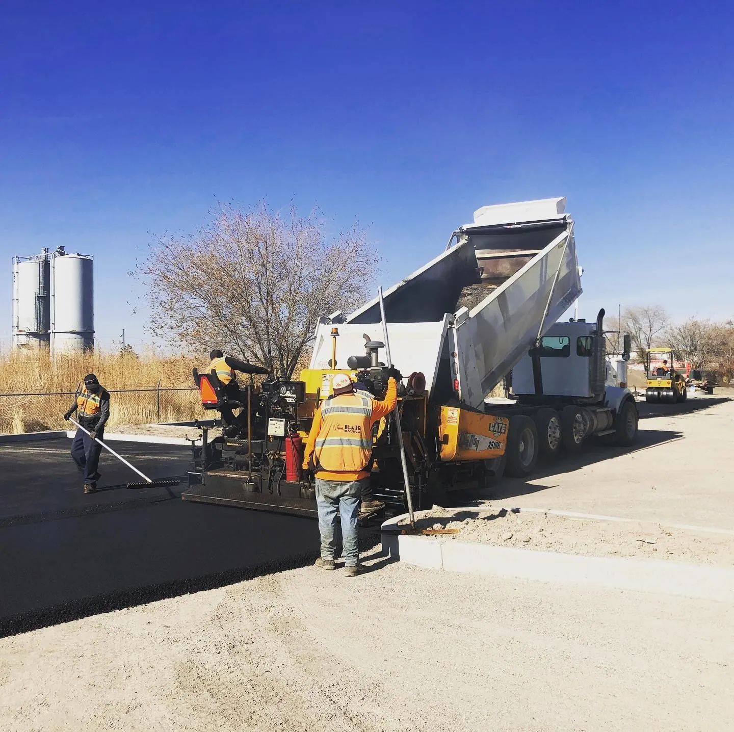 Asphalt paving in progress: truck dumping load into a paver, workers leveling the surface, clear sky.