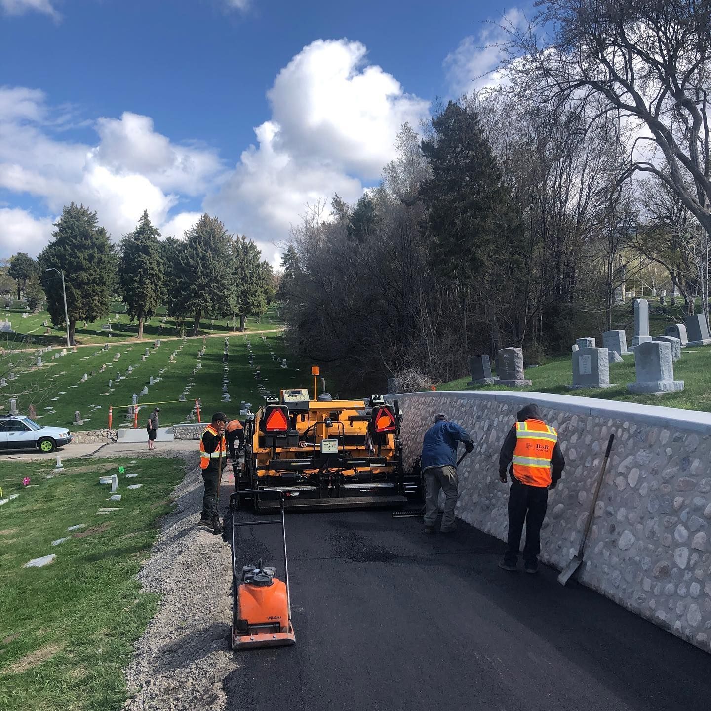 Road paving in a cemetery, asphalt being laid by workers. Trees and headstones in the background. Blue sky, bright day.