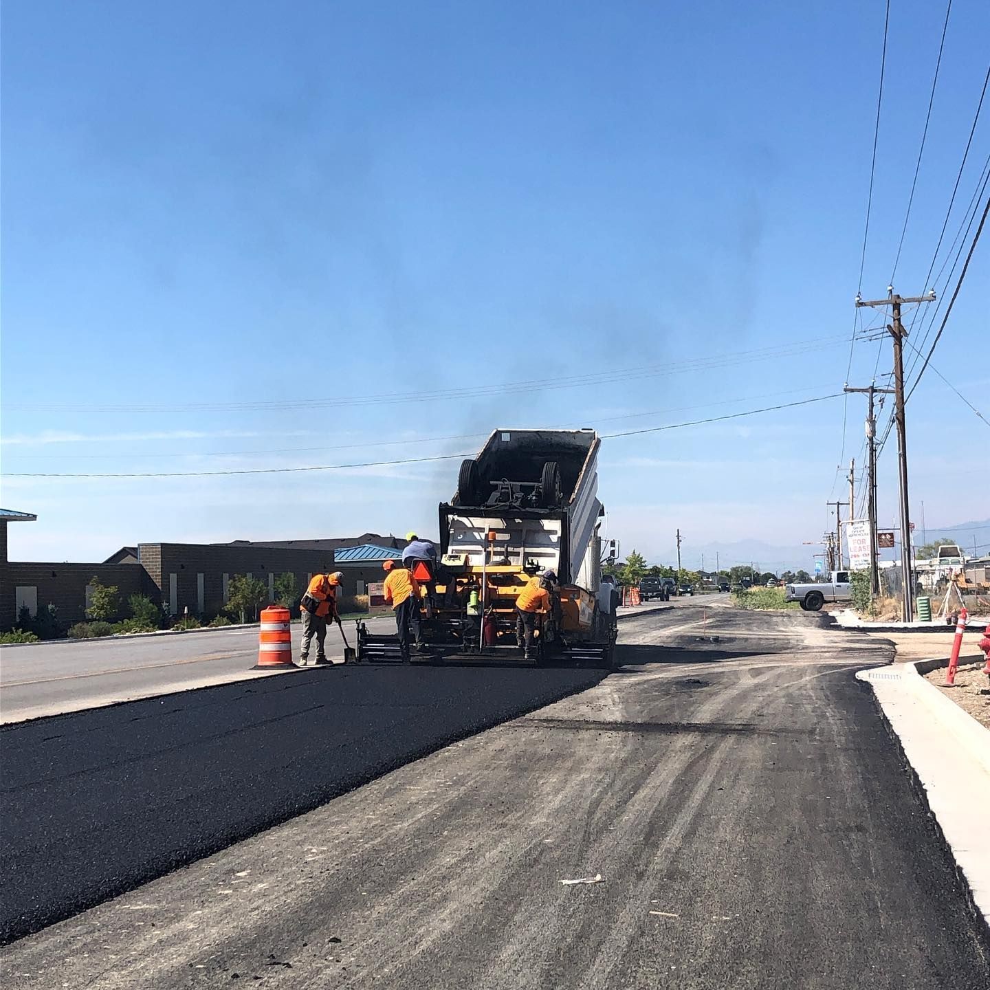 Road paving in progress; asphalt being laid by machine, workers in orange vests.