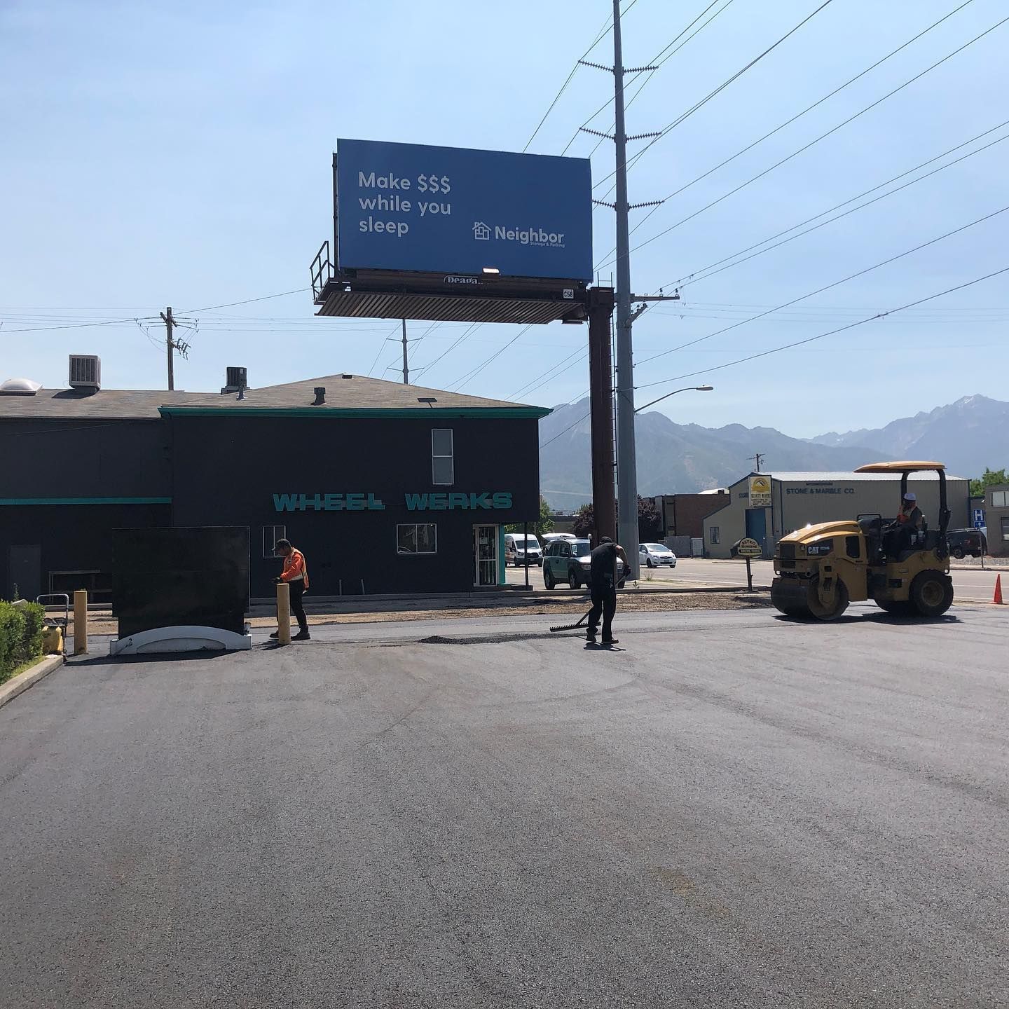 New asphalt paving in front of a building with a billboard. Workers and a roller machine are present.