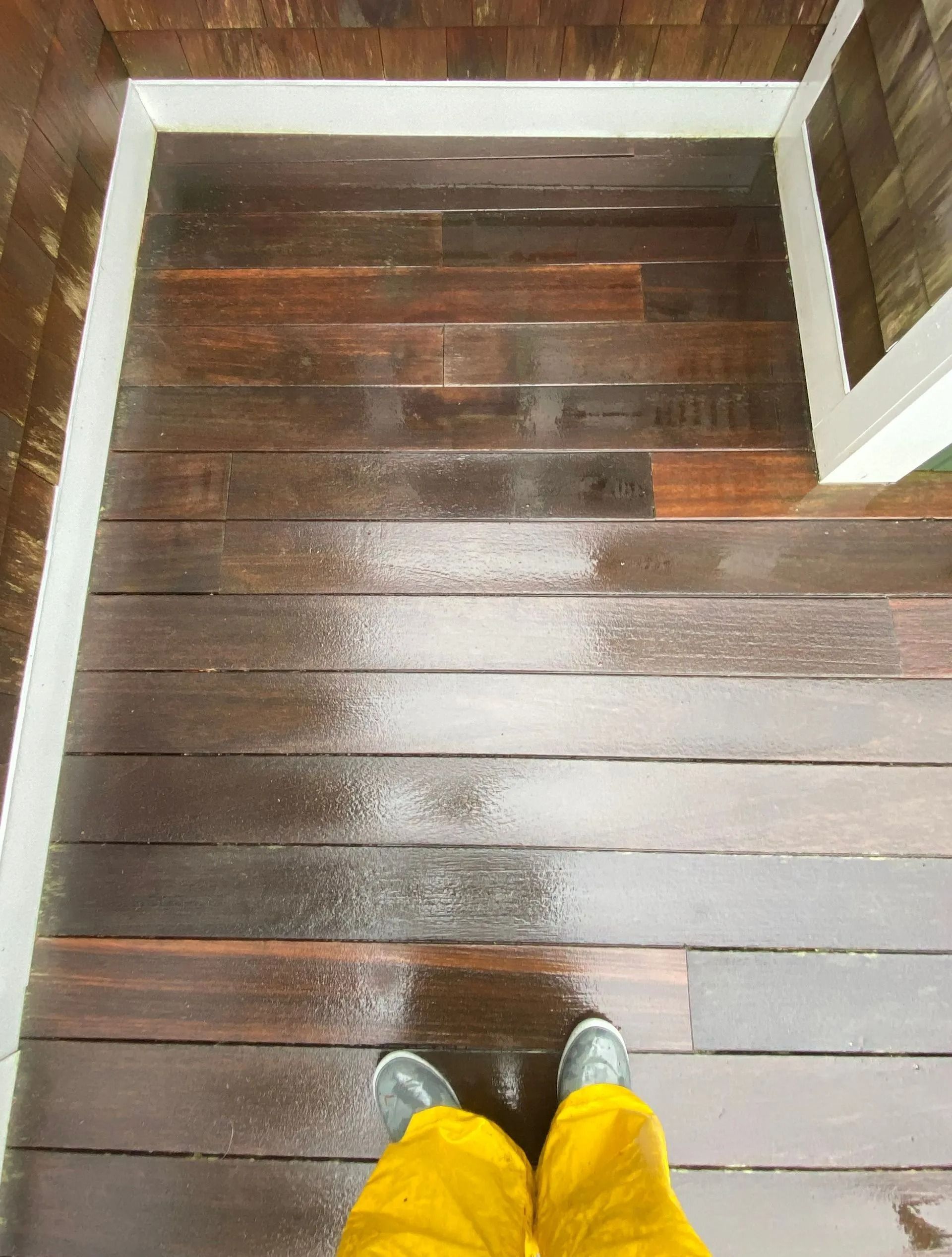 Person in yellow pants and black boots standing on a wet, dark wooden floor with white trim.