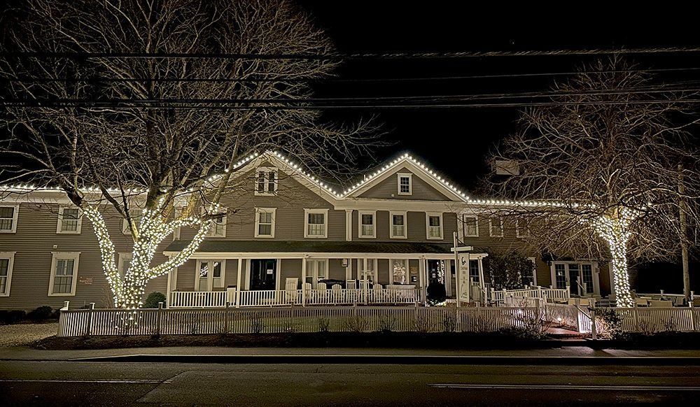 A two-story house at night decorated with Christmas lights. The trees and roof are illuminated.