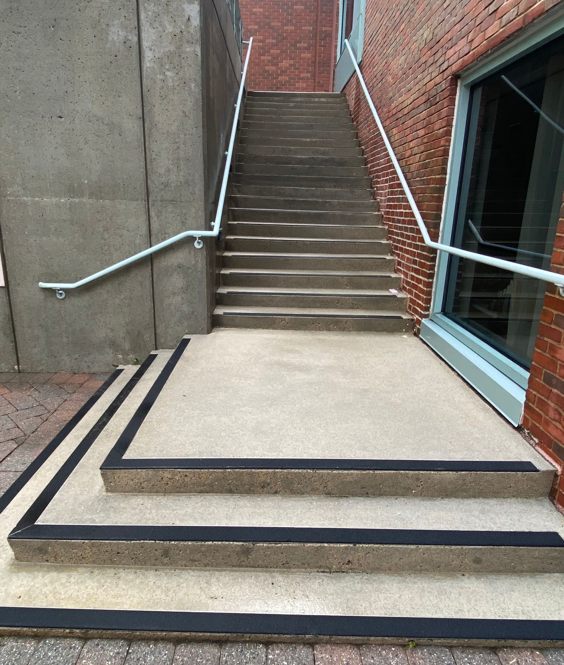 Staircase with handrails between brick and concrete walls. Black strips on the steps for safety.