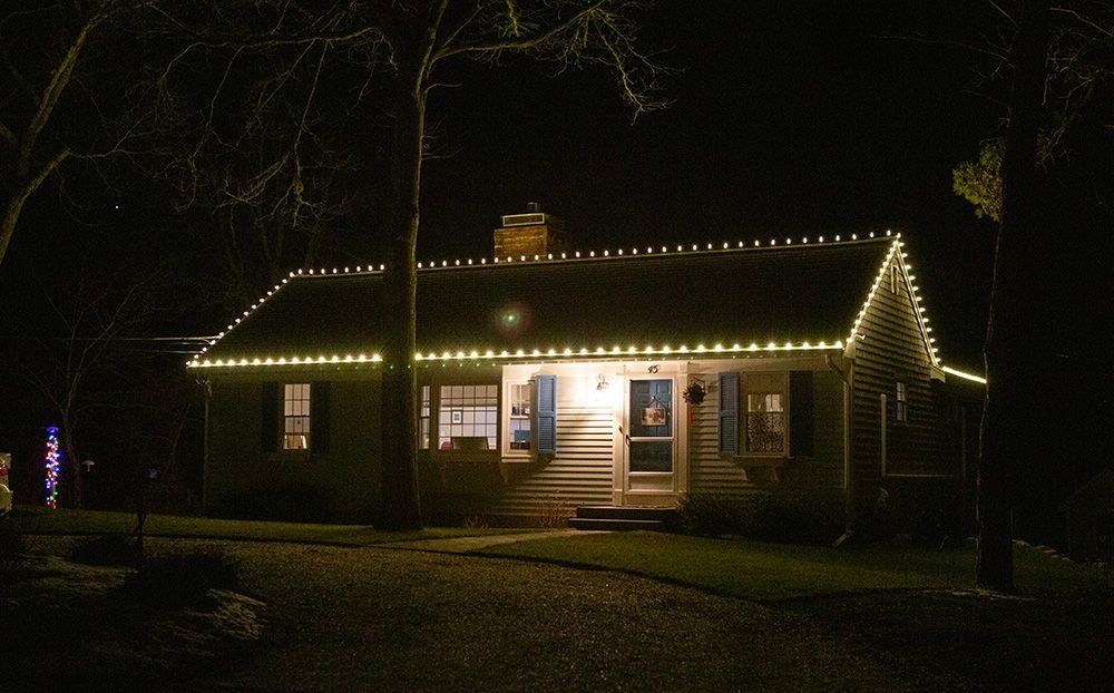 Cottage at night with warm white Christmas lights lining the roof. Lit porch, windows. Dark surroundings.