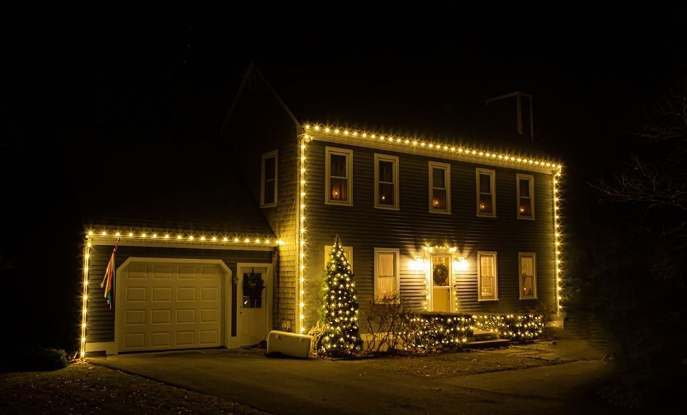 House at night, lit with warm white Christmas lights. Garage door on the left, a decorated tree on the porch.