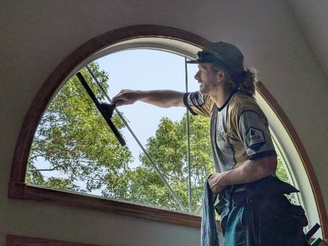 Man cleaning a curved window with a squeegee, trees visible outside.