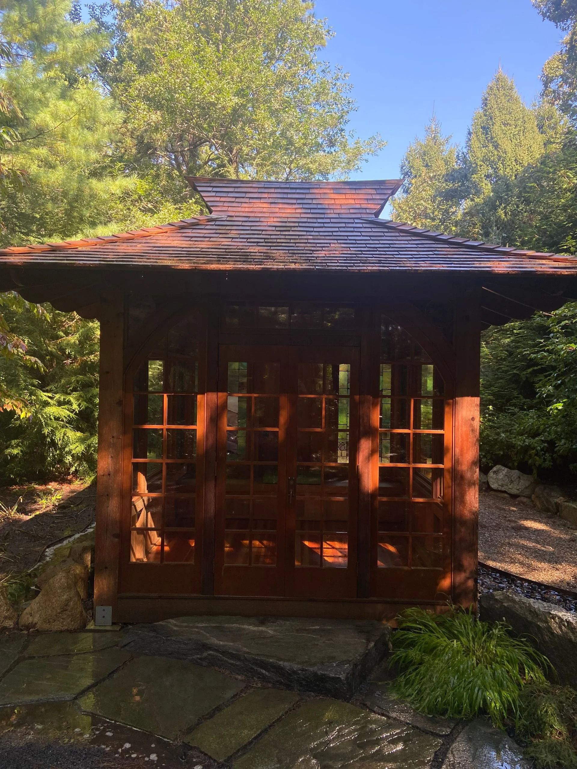 Wooden Japanese-style structure with glass doors, surrounded by lush greenery in a garden setting.