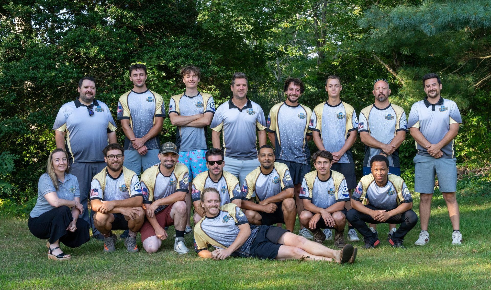 Group of 17 people in matching jerseys, posing on grass outdoors. Men and one woman, some kneeling, standing.