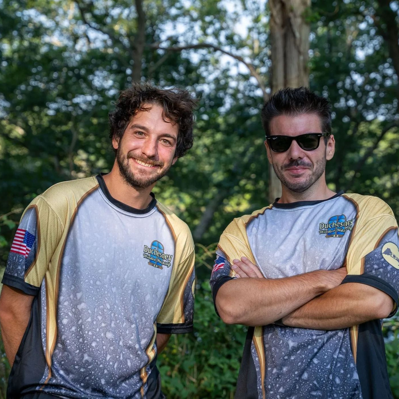 Two men in matching jerseys, smiling, pose outdoors. One has arms crossed, the other has hands at his side.
