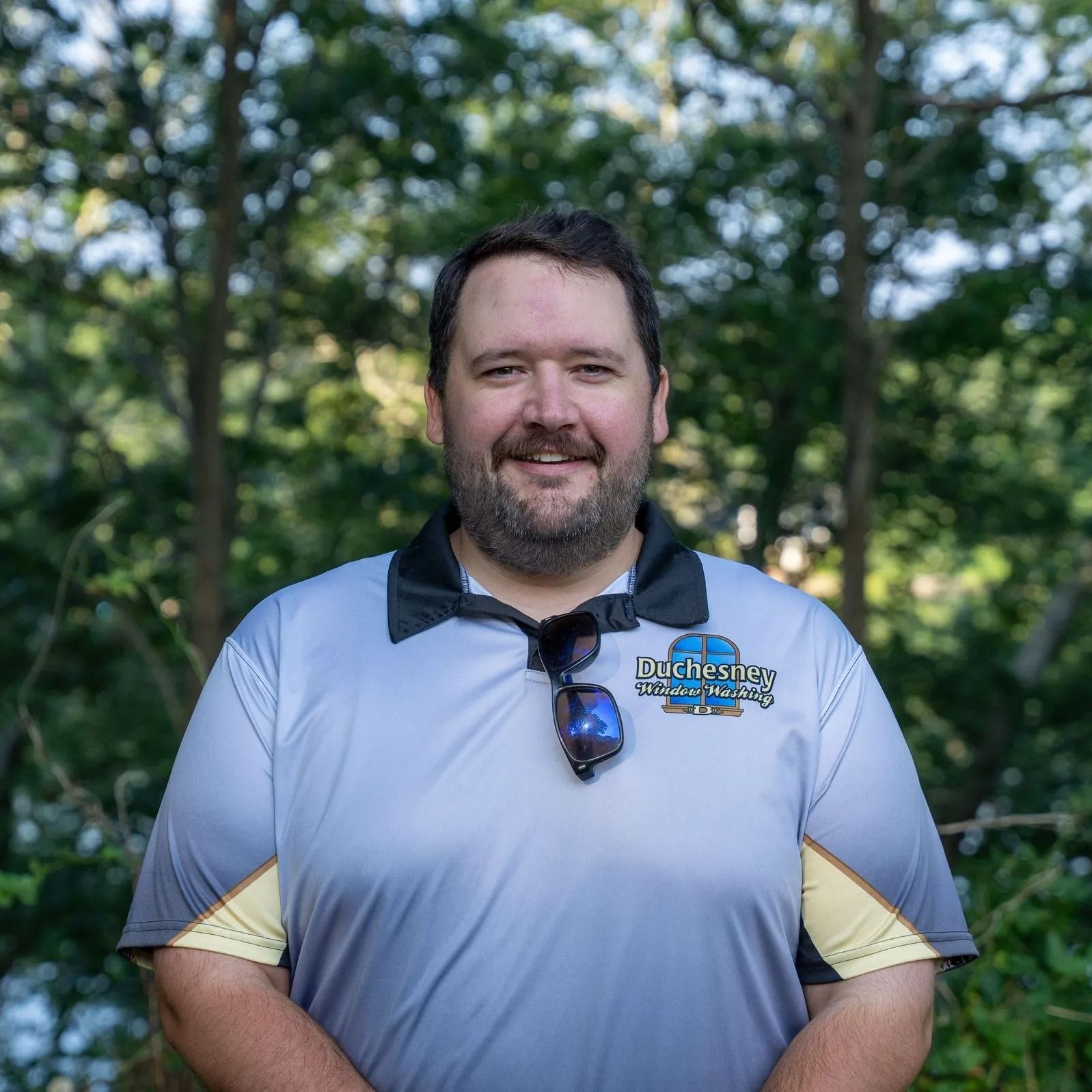 Man with beard smiles wearing a light gray polo shirt, outdoors, trees in the background.