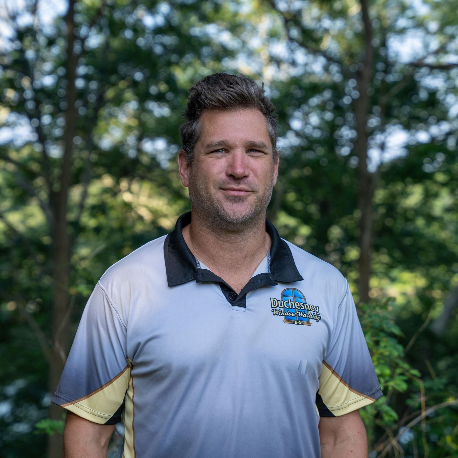 Man in gray shirt with logo smiles outdoors, trees in background.
