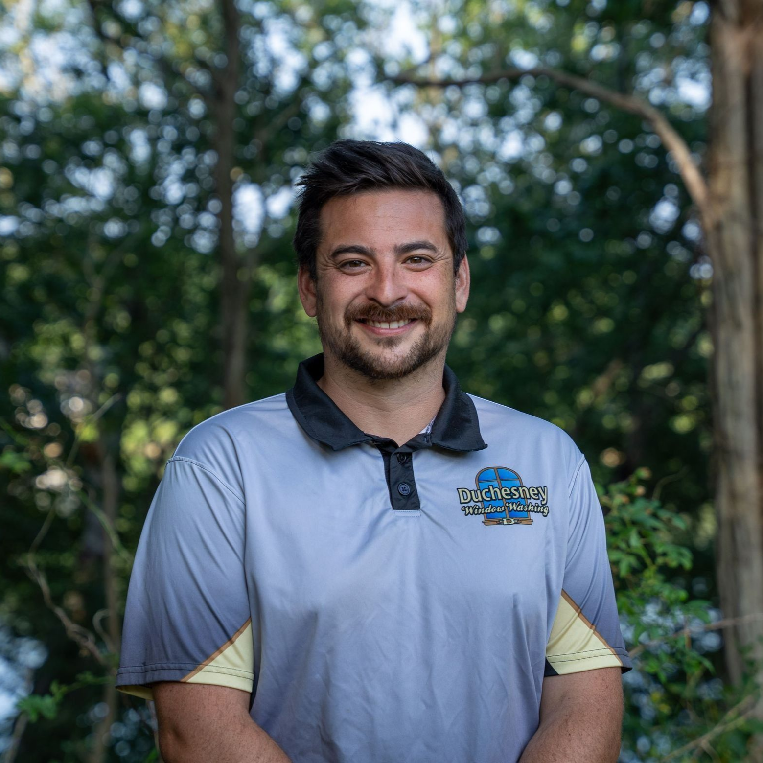 Man with a mustache smiles, wearing a grey polo shirt with a logo, outdoors, trees in background.