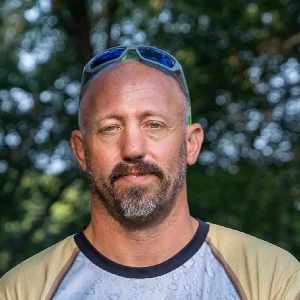 Man with shaved head, sunglasses on forehead, outdoors, looking at the camera.