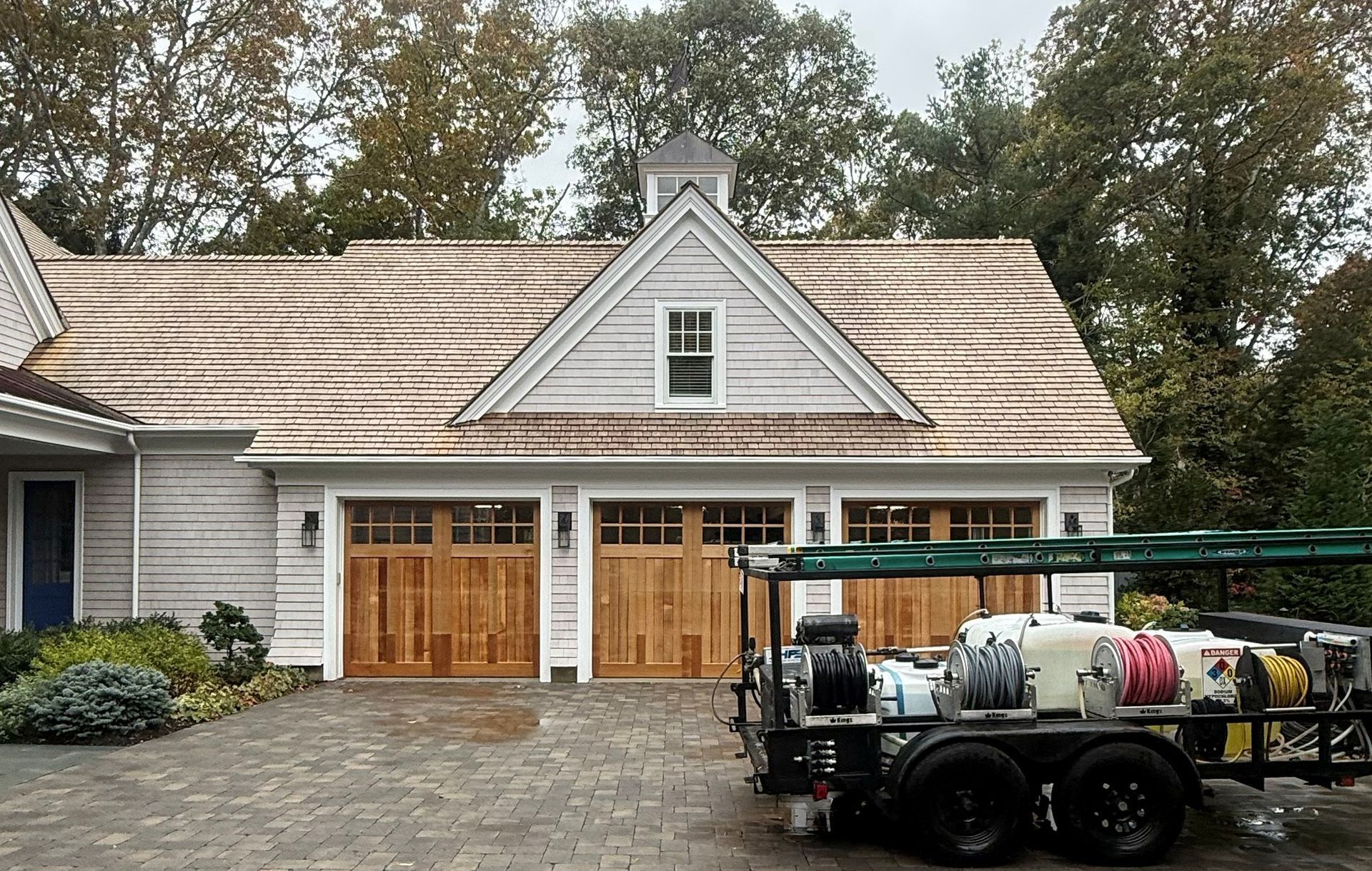 Garage with wooden doors, gray siding, and a trailer with cleaning equipment parked on a brick driveway.