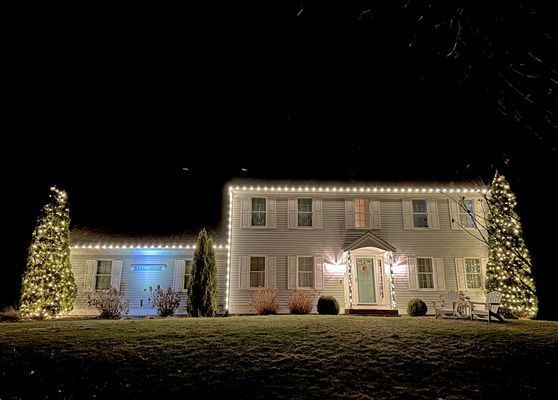 House decorated with Christmas lights at night. White lights outline roof, with trees on either side.