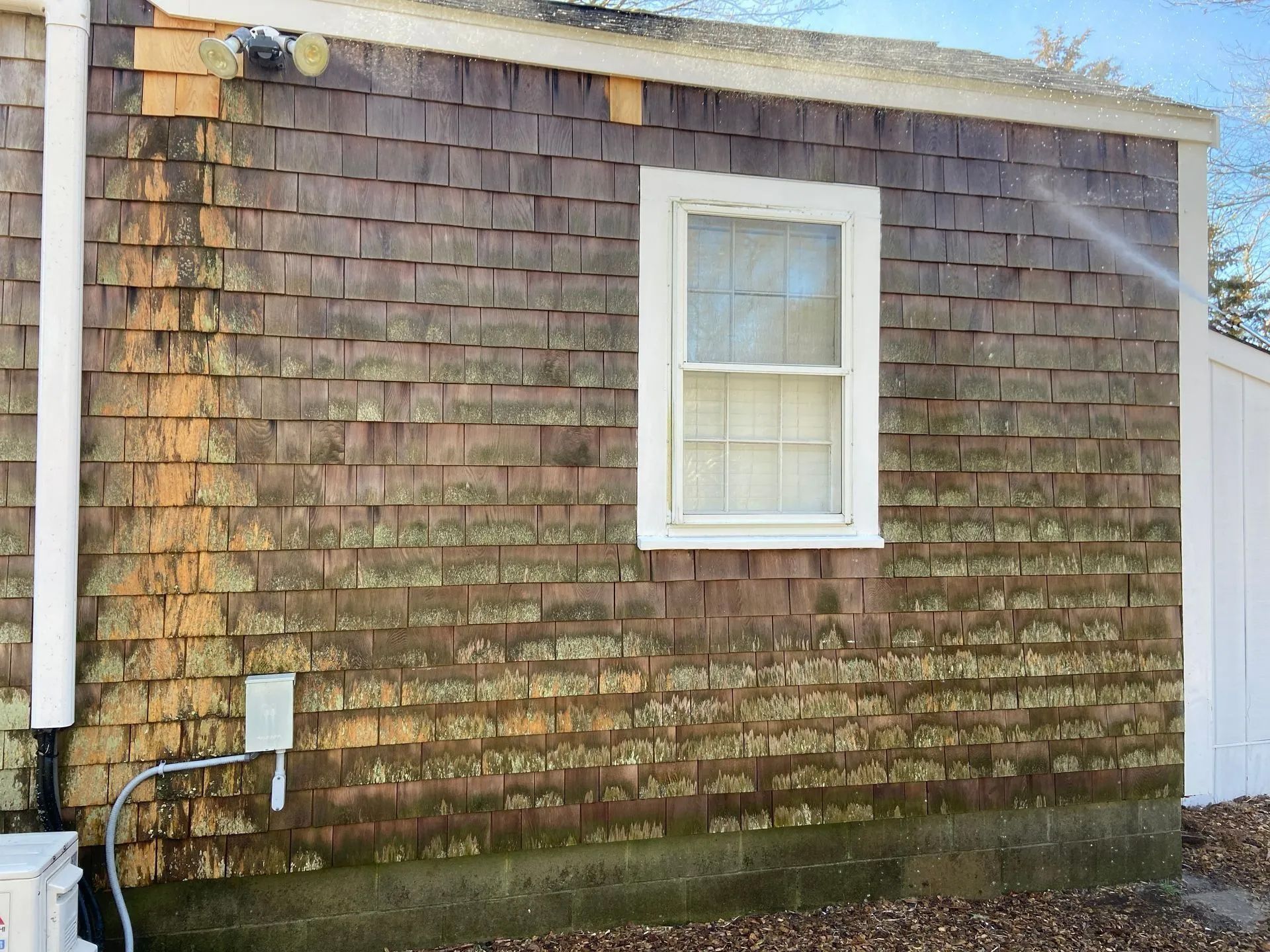 Pressure washing a weathered, brown shingle siding on a house; window visible.