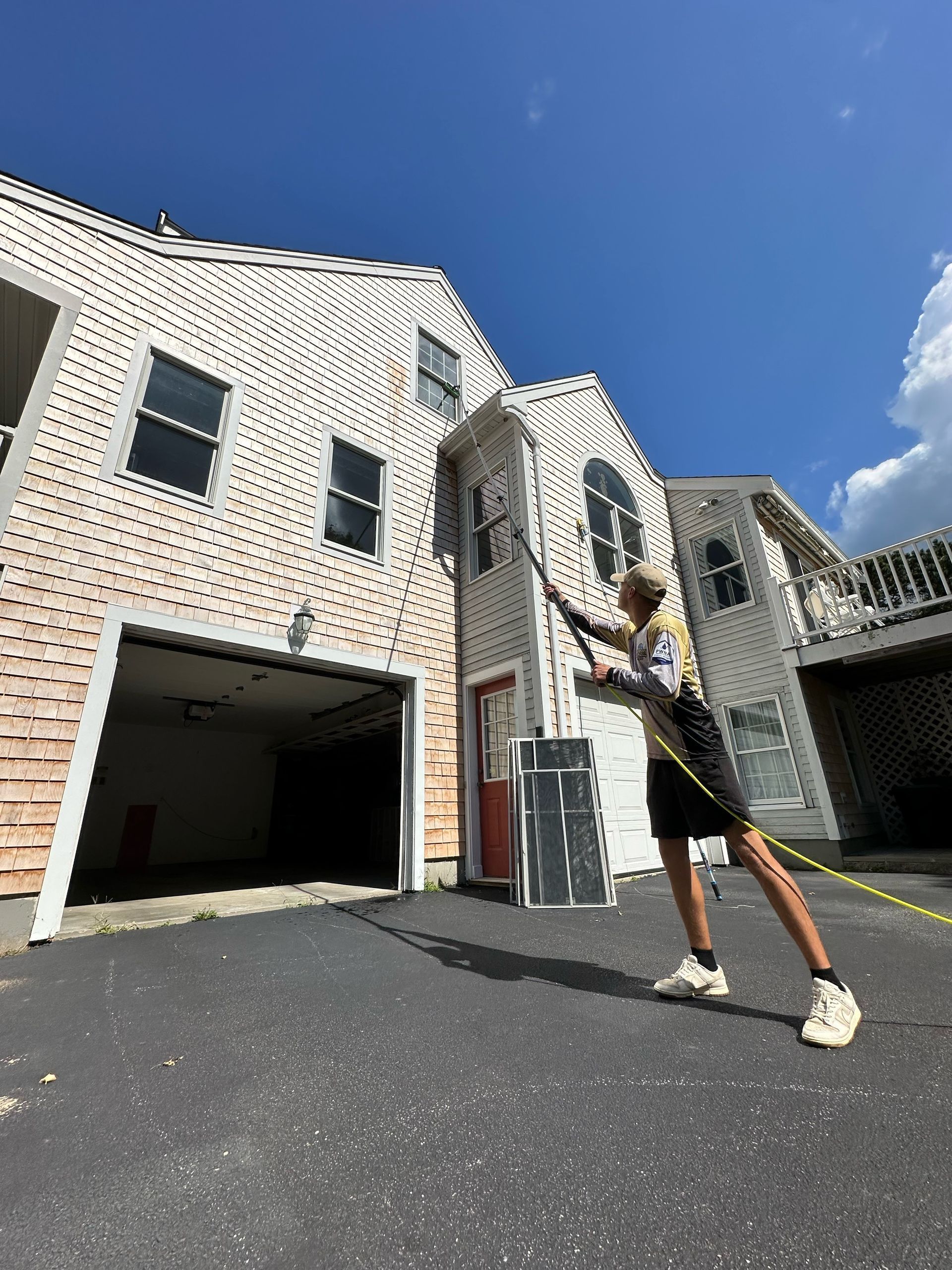 Person power washing a two-story white house with an open garage on a sunny day.